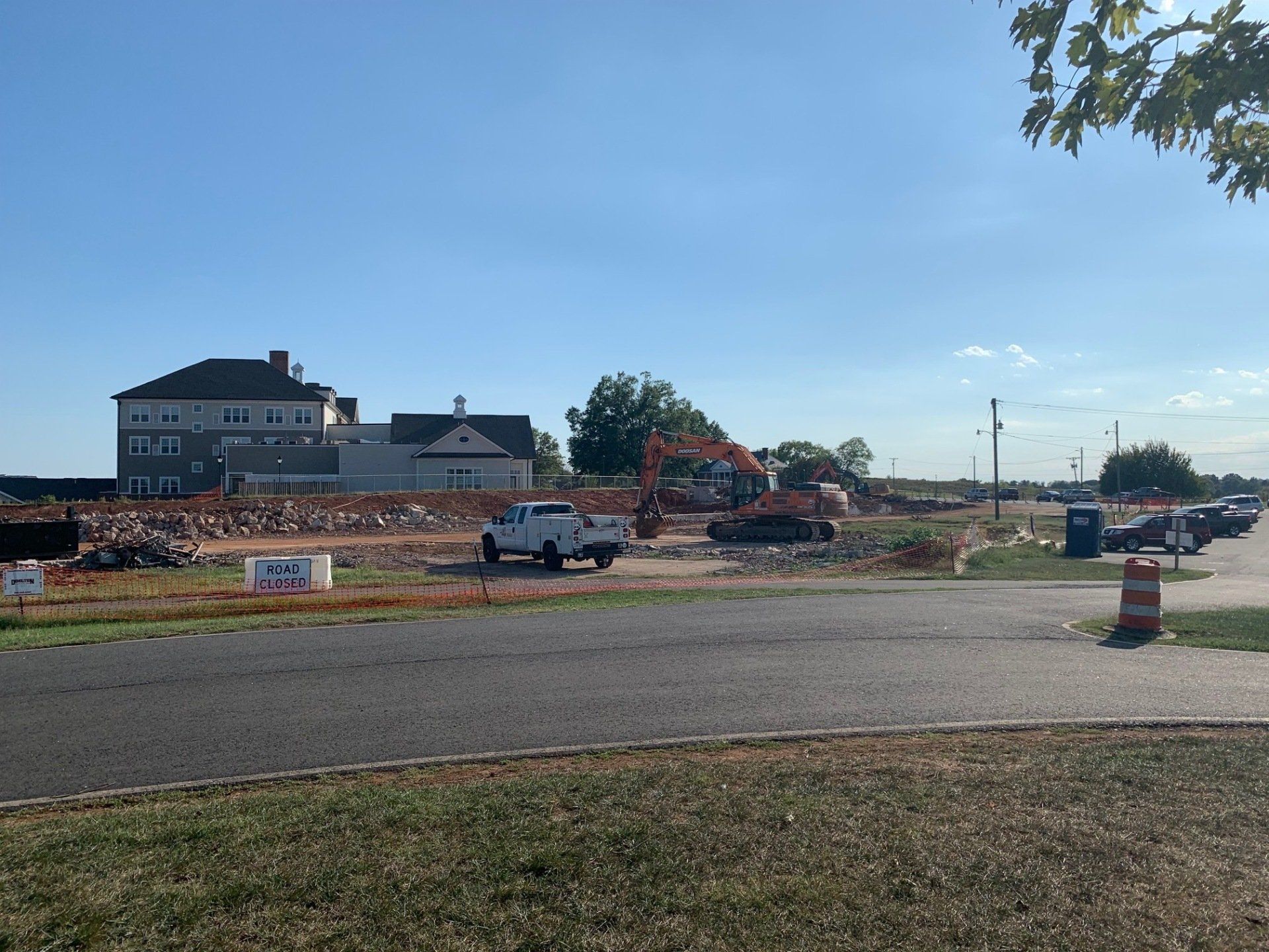 Road construction site with trucks, dirt piles, and a house under a clear blue sky.