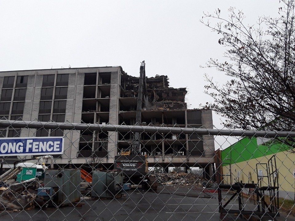 Partially demolished building behind a fence, with rubble and construction equipment under an overcast sky