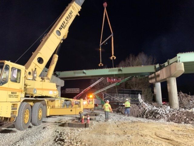 Crane lifting a bridge beam at a nighttime construction site with workers below