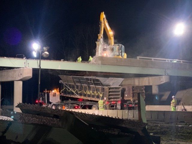 Nighttime construction site with a yellow excavator atop a concrete overpass under bright lights
