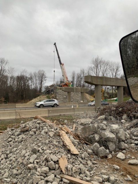 Construction site with crane lifting stone near road, rubble pile in foreground, overcast sky.