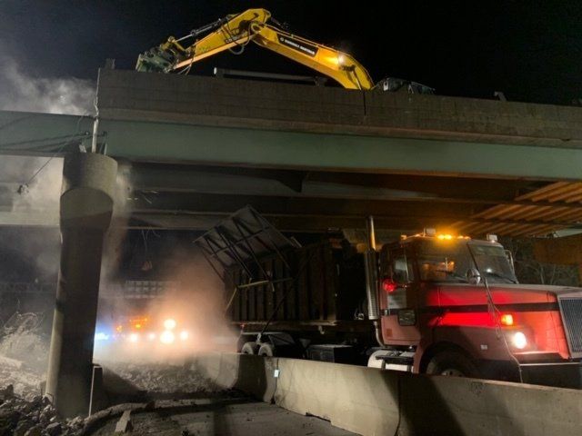 Yellow excavator and red truck working under an overpass at night, with dust and headlights visible