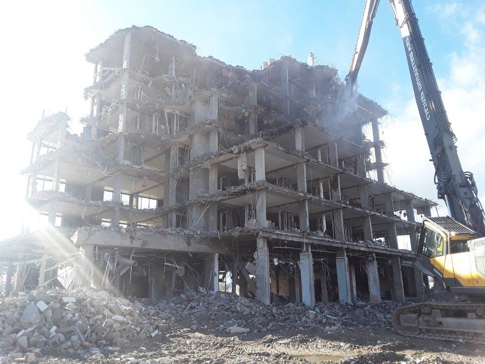Demolished concrete building with rubble in front and an excavator beside it