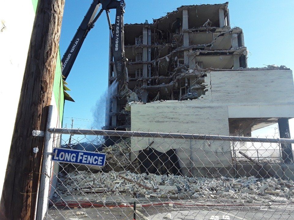 Demolition of a damaged building behind a chain-link fence with rubble and a crane arm visible