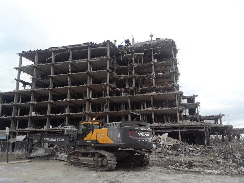 Tracked excavator amid rubble before a damaged multi-story building under demolition