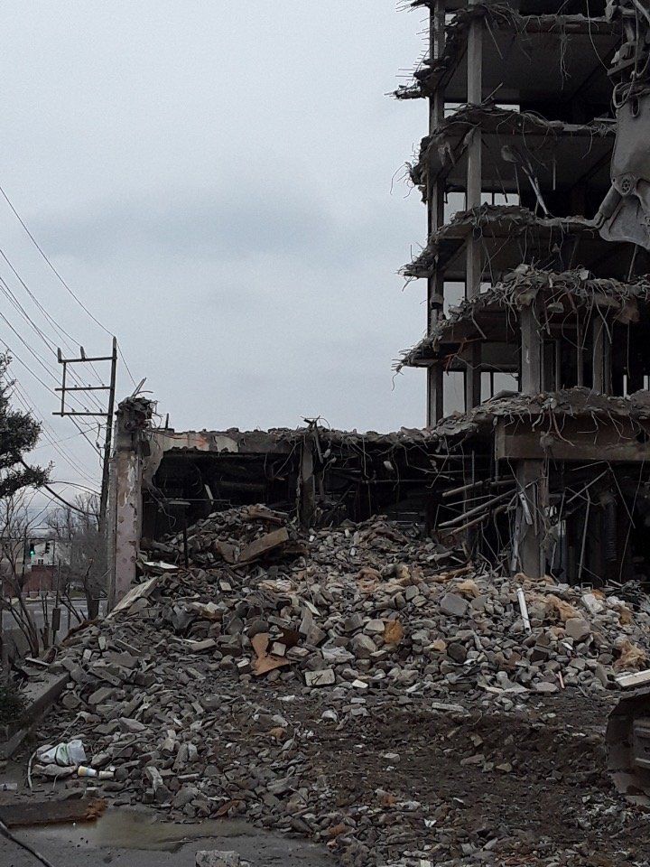 Collapsed building and rubble after destruction, with a damaged multi-story structure in the background.