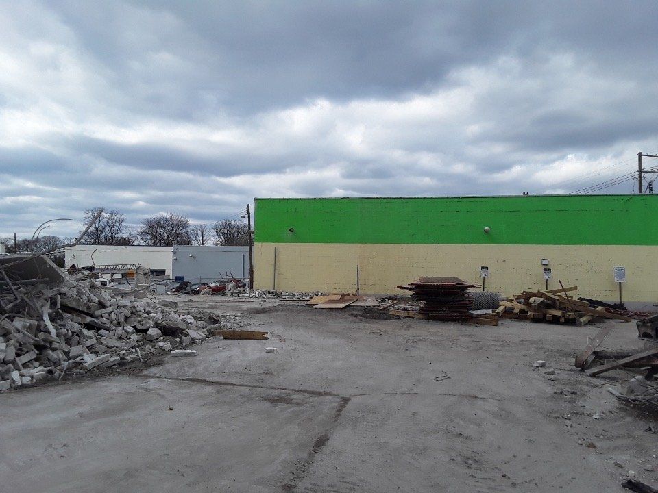 Empty industrial lot with rubble piles, broken pavement, and a green-roofed building under cloudy sky