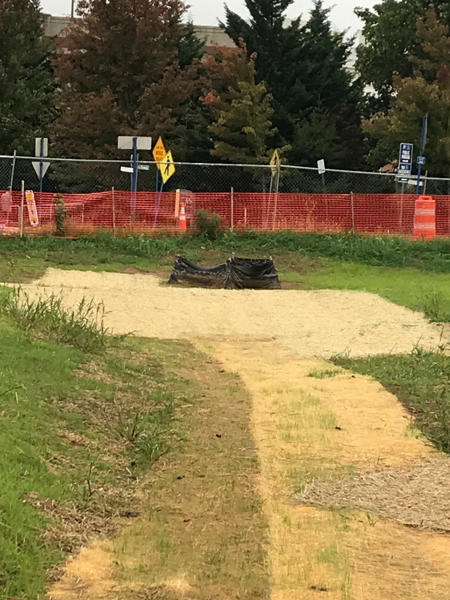 Black tarp over a dirt mound on a sandy path beside orange construction fencing and trees.