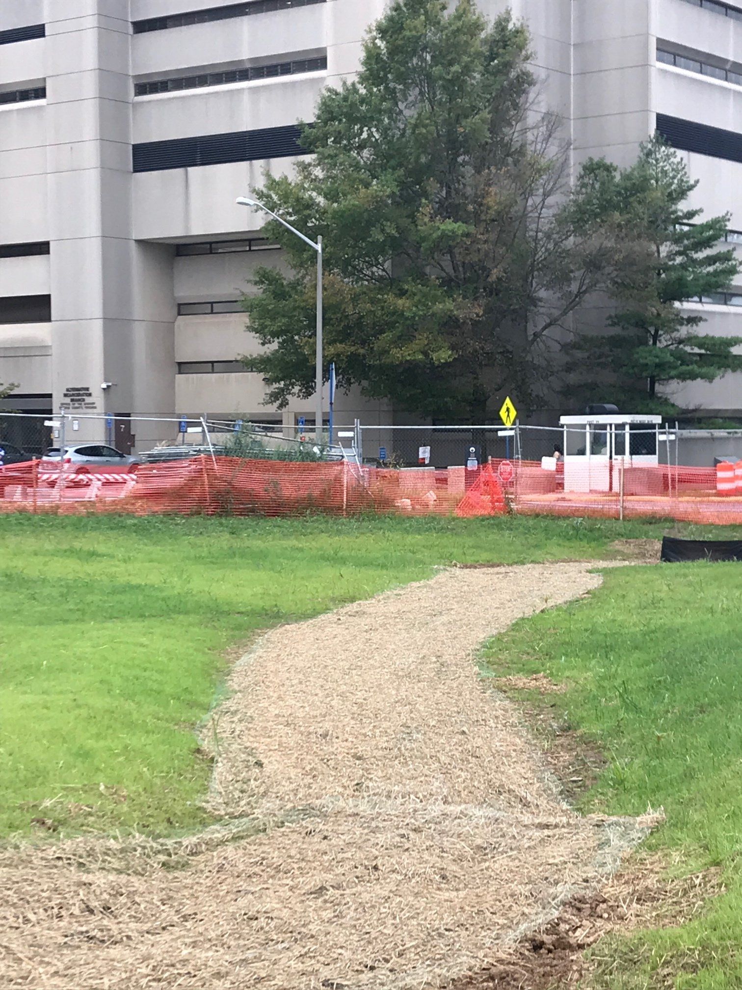 Gravel path curving through grass toward a building, with orange construction fencing and trees nearby