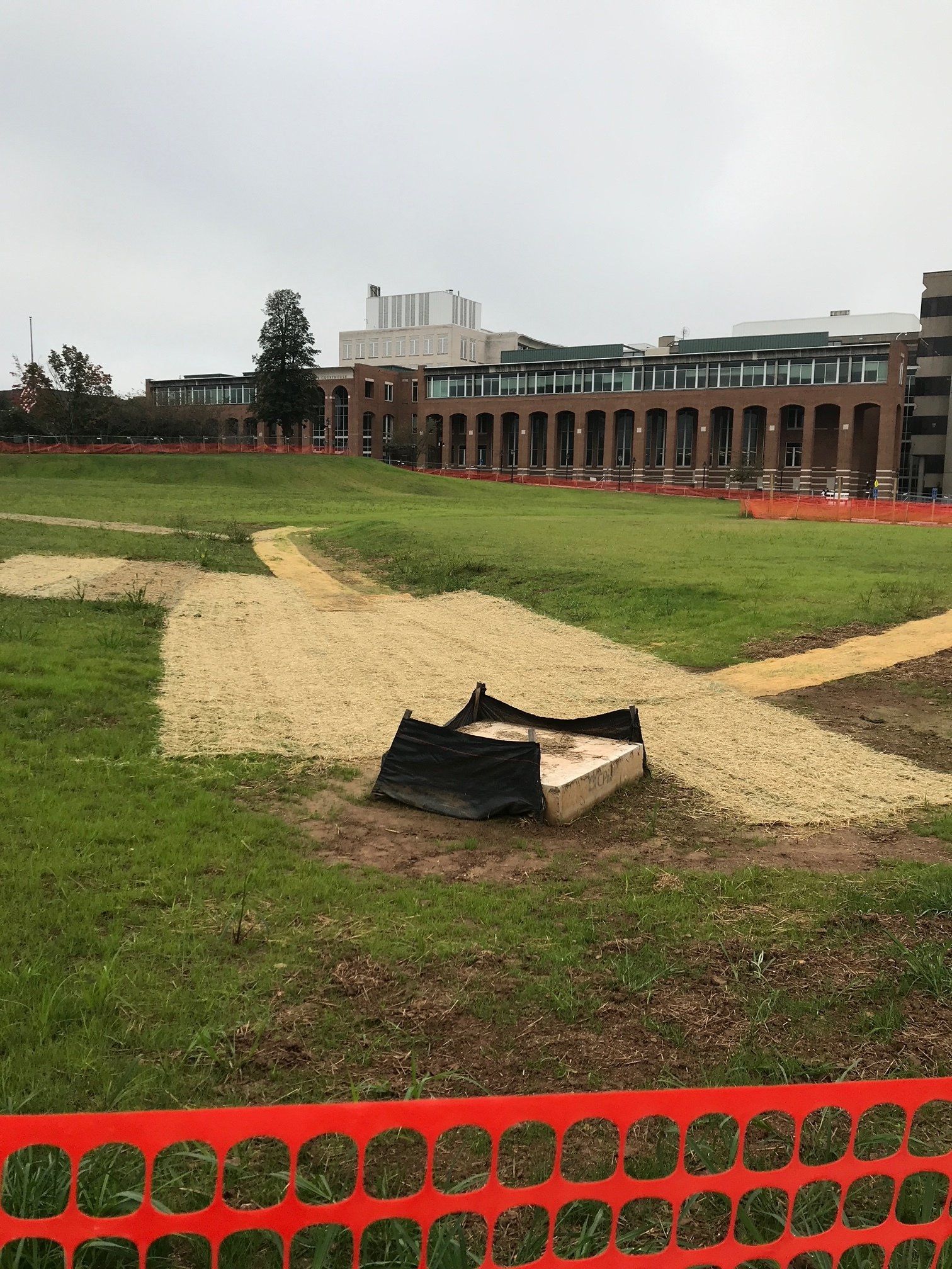 Grassy field with dirt paths and a brick building in the background, partly fenced with orange netting