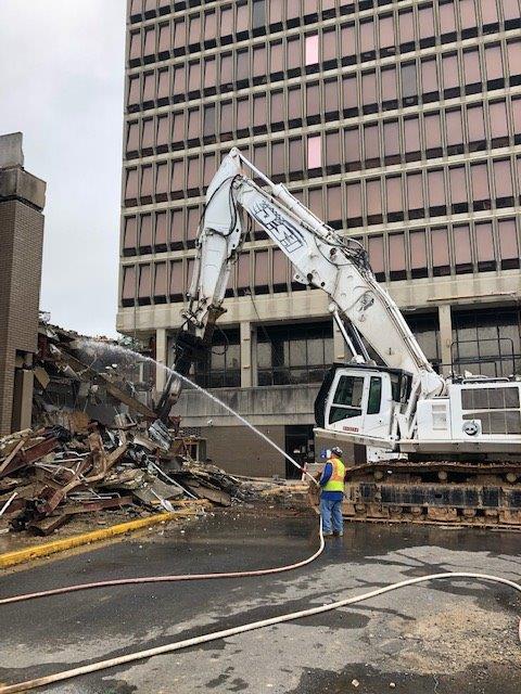 White excavator demolishing rubble beside a tall office building, with a worker in a blue vest nearby.