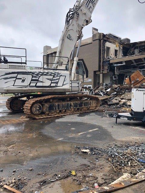Excavator amid rubble at a demolished building site, with smoke and debris on the ground.
