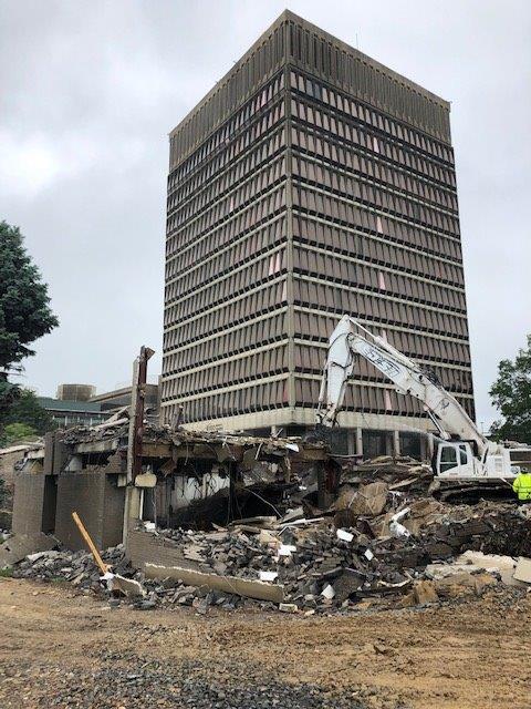 Demolition excavator clearing rubble beside a damaged high-rise building under a cloudy sky