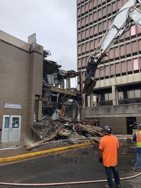 Demolished building facade with an excavator clearing rubble on a city street; two workers stand nearby.