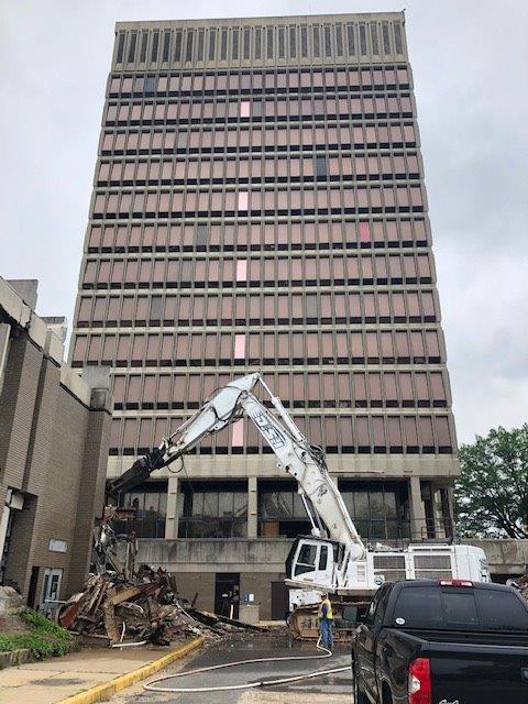 Demolition excavator in front of a tall office building, with debris and a parked pickup truck.