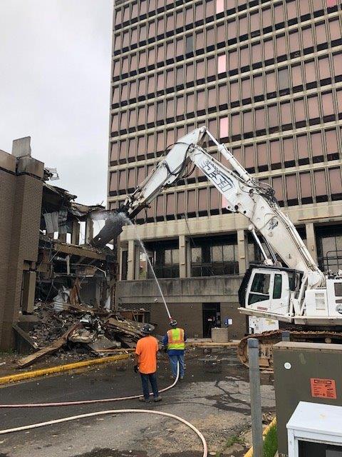 Demolition scene with excavator tearing down a building beside a tall office tower; two workers nearby