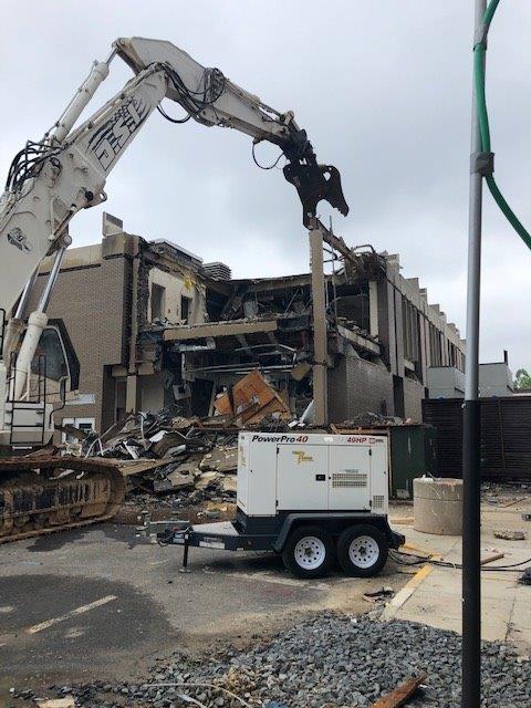 Excavator demolishing a damaged building with rubble and a portable generator on site.