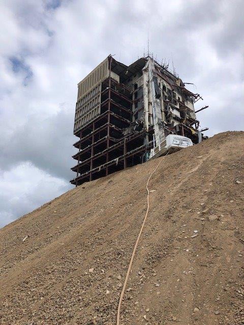 Damaged high-rise on a steep dirt slope under cloudy sky, with a cable running down the hill