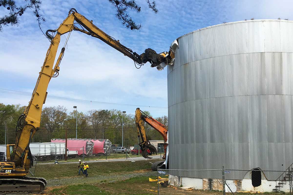Excavator demolishing a large silver metal tank at an outdoor work site
