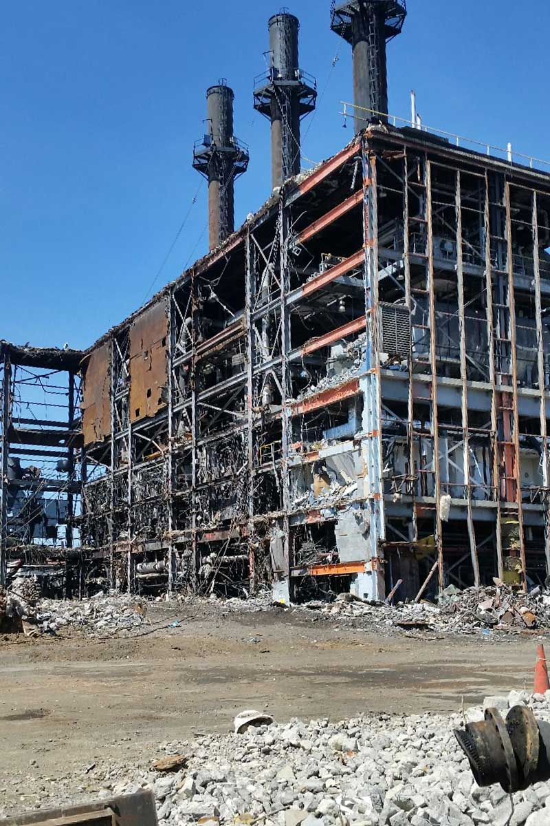 Charred industrial building skeleton after fire, with smokestacks and debris under a clear blue sky