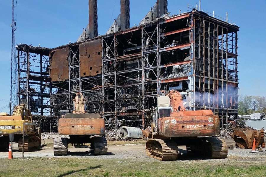 Partially burned building with excavators and dump trucks in front, under a clear blue sky