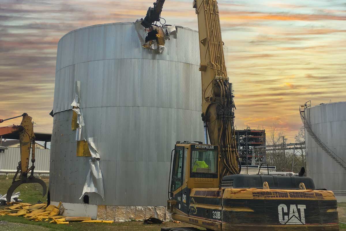 Excavators dismantling large cylindrical metal storage tanks at sunset, with a CAT machine in the foreground