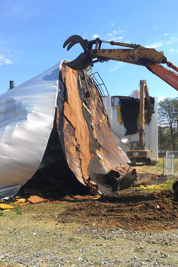 Excavator tearing down a damaged metal building, with twisted siding and debris on the ground.