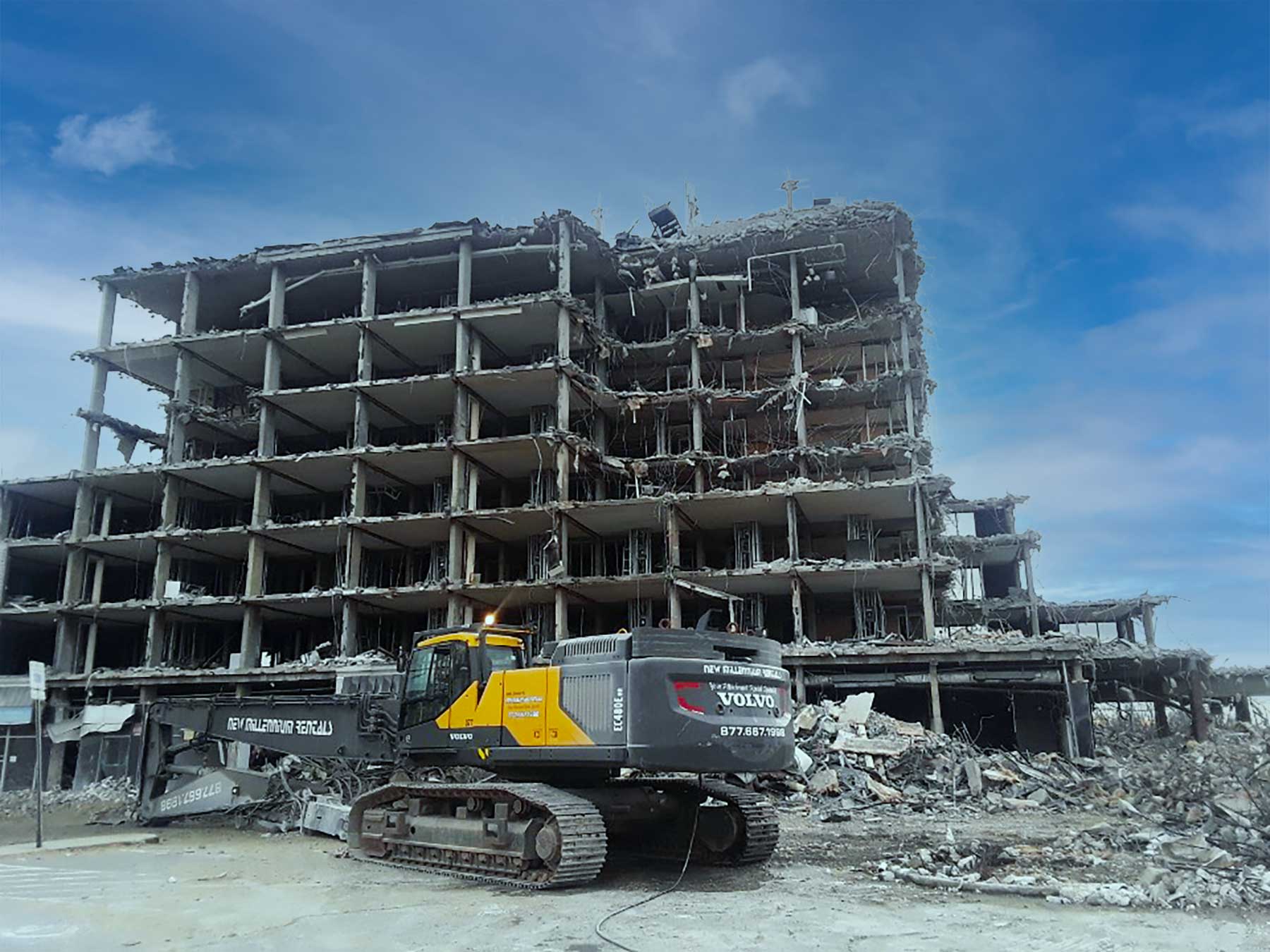 Excavator in front of a partially demolished multi-story building under a blue sky