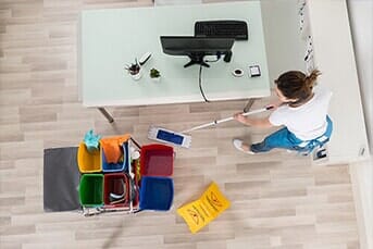 Cleaning person mopping office floor near supplies and a desk with a computer.