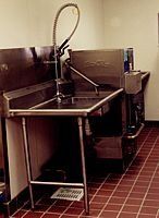 Industrial stainless-steel utility sink with faucet in a tiled room, beside a metal machine.