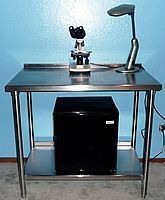 Black laboratory workstation with a microscope, task lamp, and mini fridge on a metal table