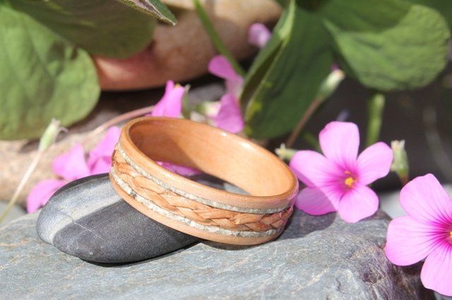 Wooden ring with braided inlay and ashes resting on a small stone and surrounded by tiny pink flowers.