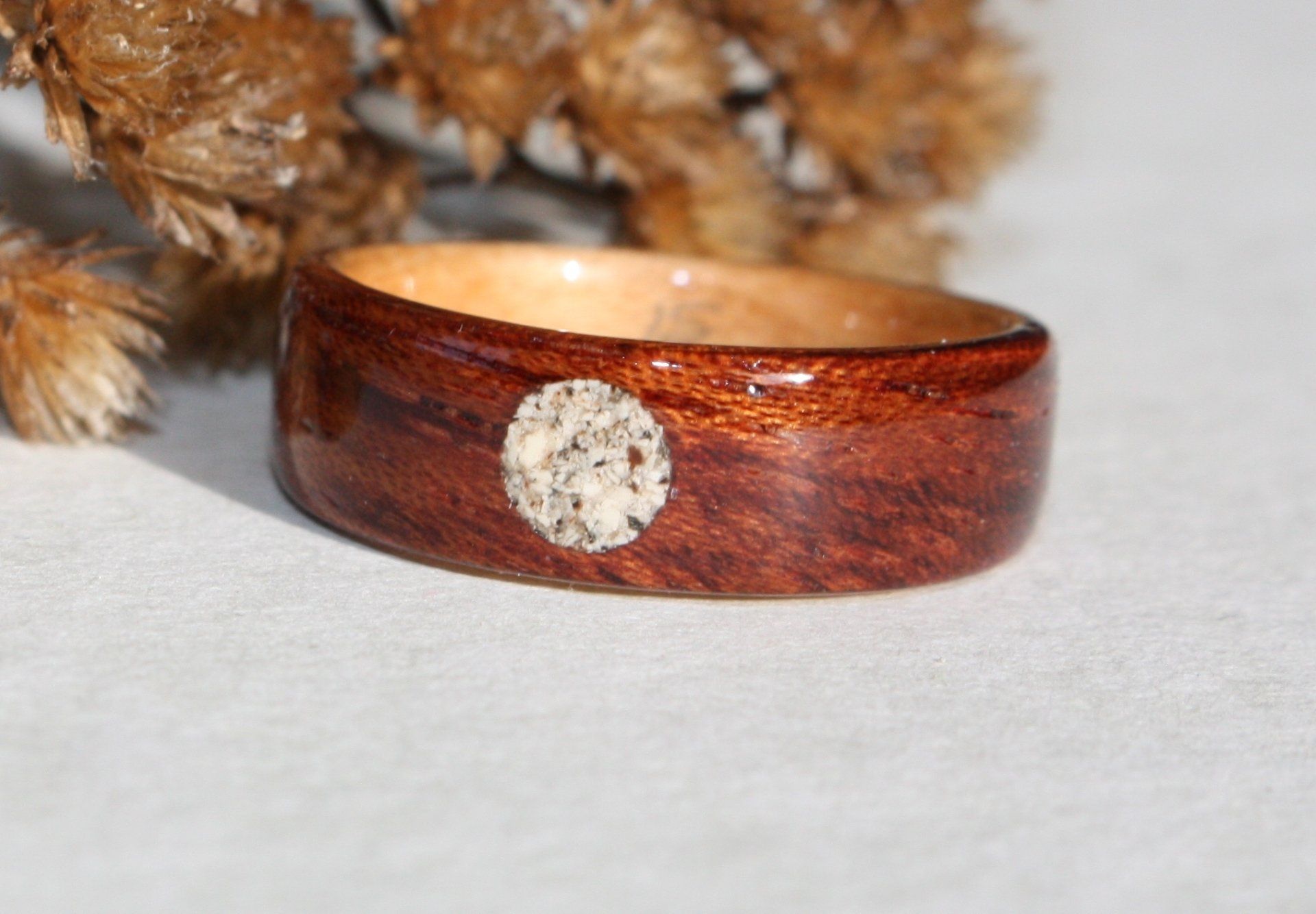 Wooden memorial ring with a round inlay of ashes. Behind the ring are dried flowers.