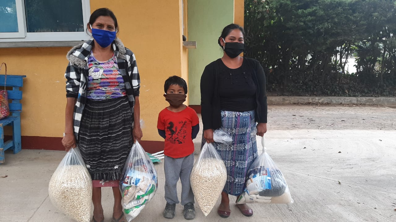 Drei Menschen mit Masken stehen nebeneinander und halten Tüten mit Lebensmitteln.