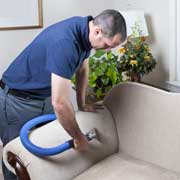 A professional home cleaner uses a vacuum on a couch arm.