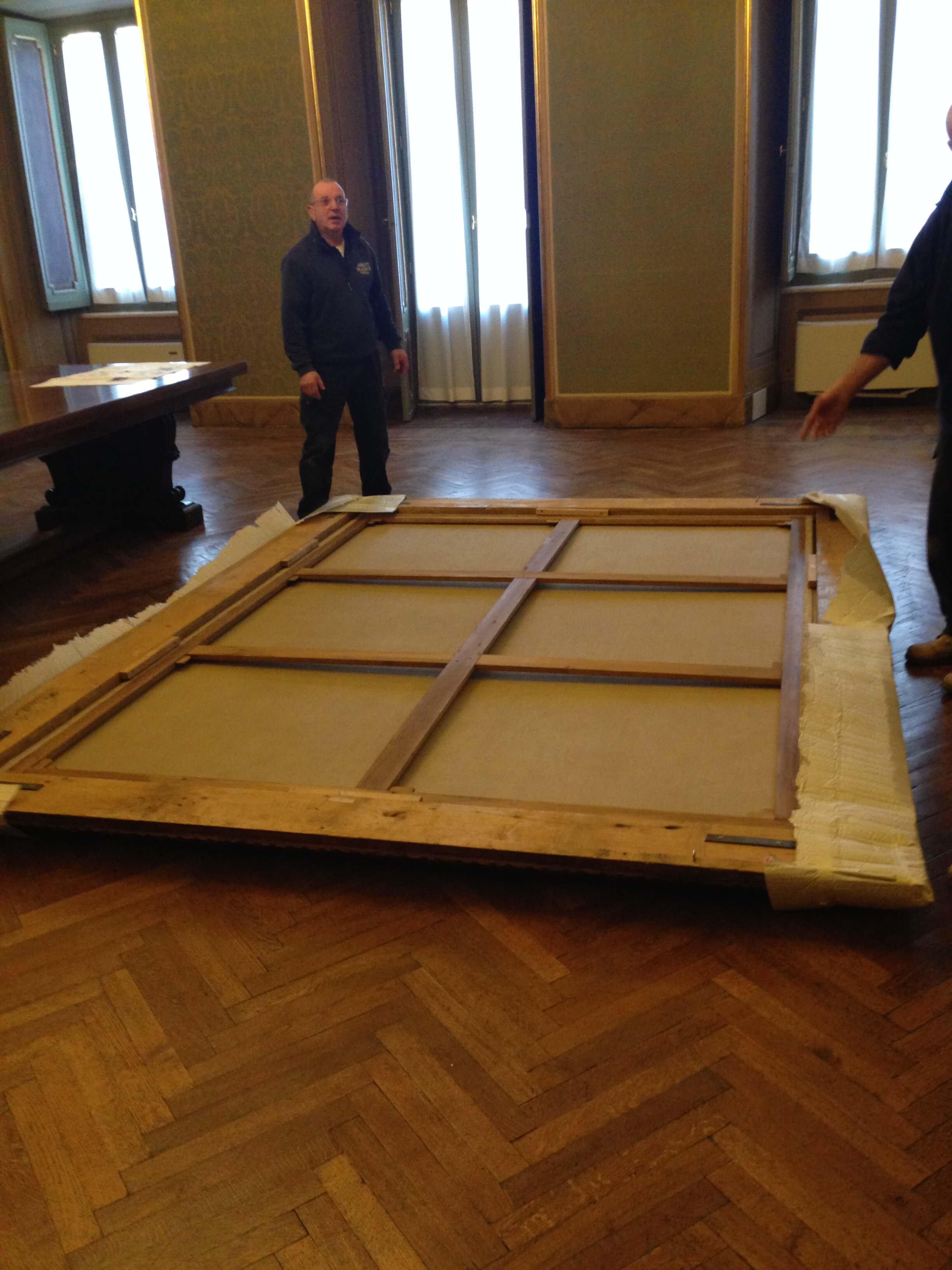Grande opera d'arte, fissata a un pallet di legno, all'interno. Due persone vicine; pavimento in legno, finestre alte.