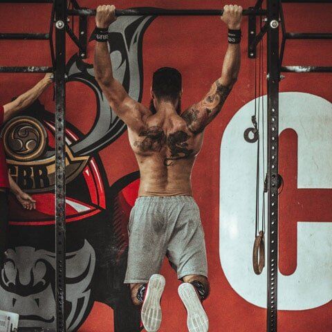 A shirtless person with back tattoos performs a pull-up on a metal gym rig against a red wall with graphic designs.