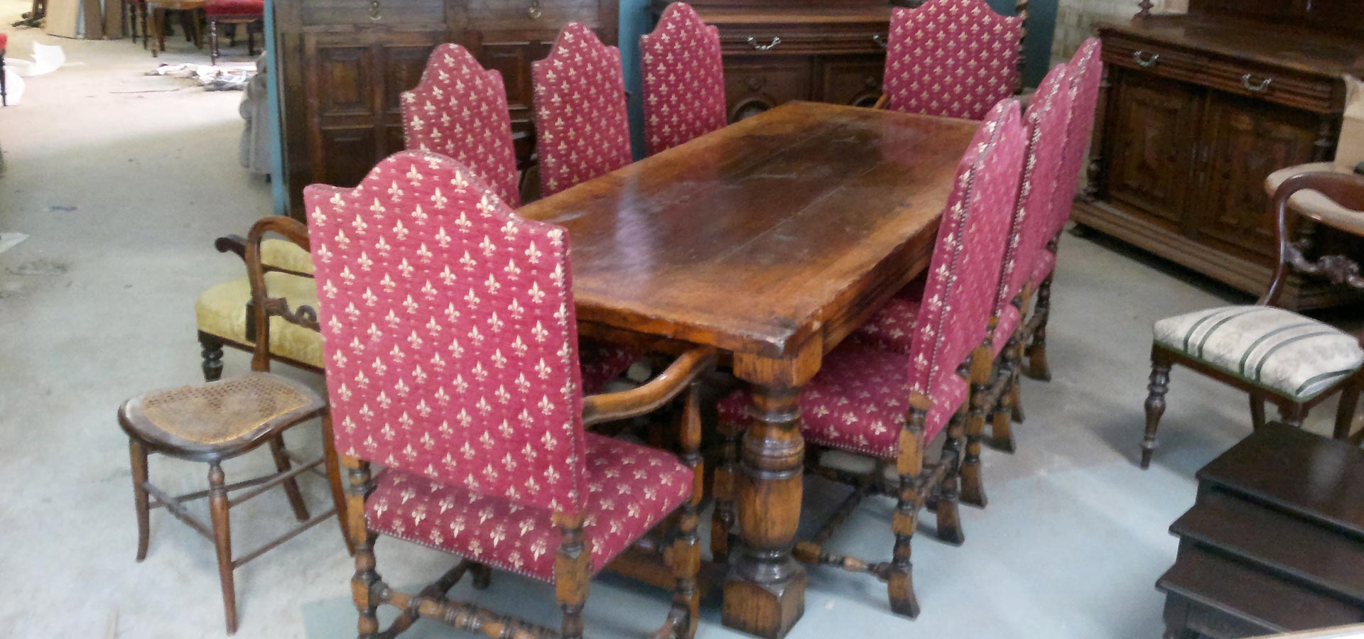 A wooden dining table with ornate legs, surrounded by red upholstered chairs in a warehouse setting.