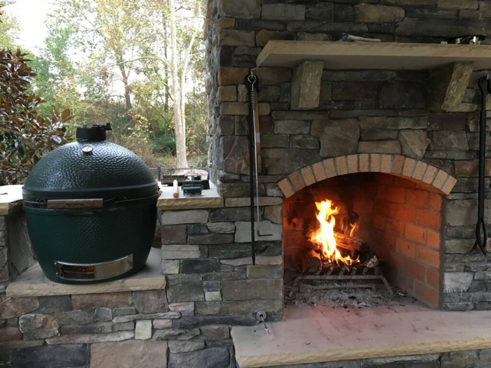 Patio with stone fireplace and burning fire beside a green ceramic grill under a covered outdoor area