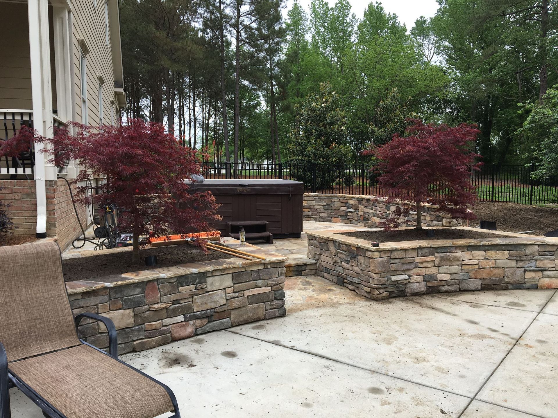 Stone patio with retaining walls, red shrubs, and a brown hot tub in a wooded backyard