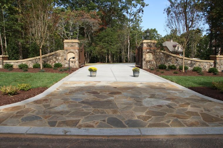 Stone driveway leading to a gated entrance with stone pillars, flanked by flower beds and trees