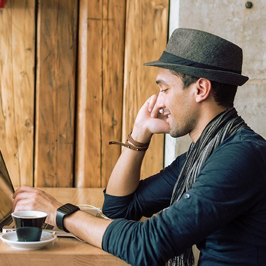 man sitting in a cafe with laptop, talking on phone, wearing a fedora hat inside
