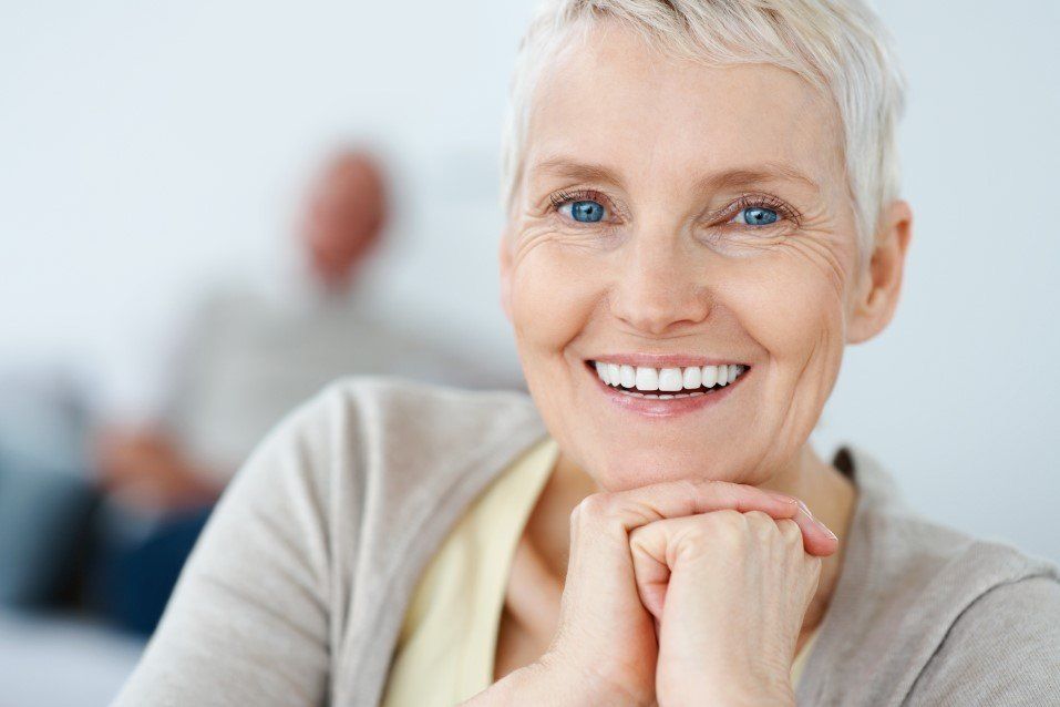 Smiling person with short white hair resting chin on hands in a bright office setting
