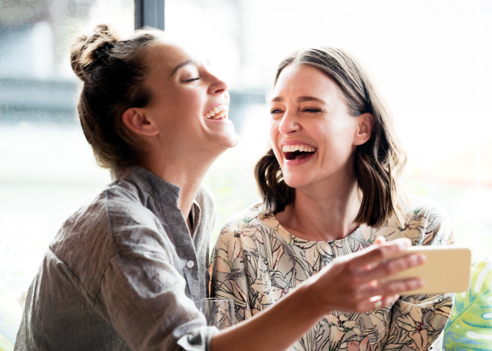 Two people laughing together indoors while one holds a phone and a cup of coffee