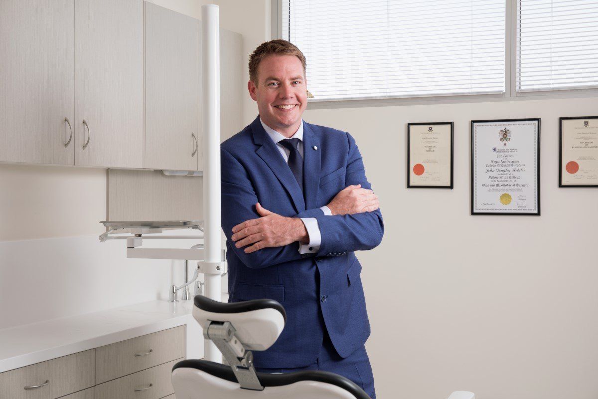 Dentist in blue suit standing in a bright exam room beside a dental chair and certificates on the wall