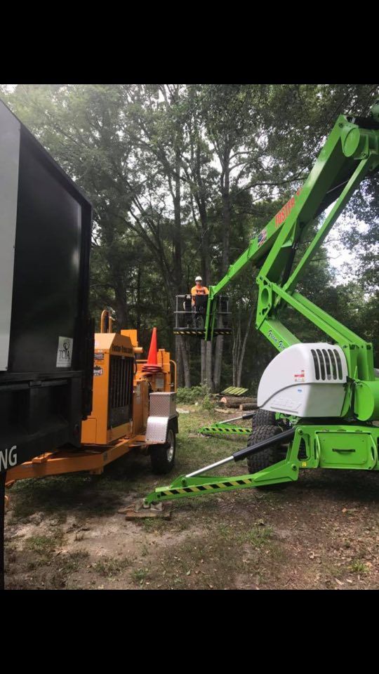 Tree service worker in lift trimming tree; wood chipper and truck present.