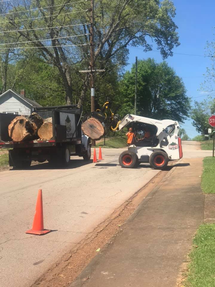 A skid steer loads logs into a dump truck on a street. A worker wearing orange directs.