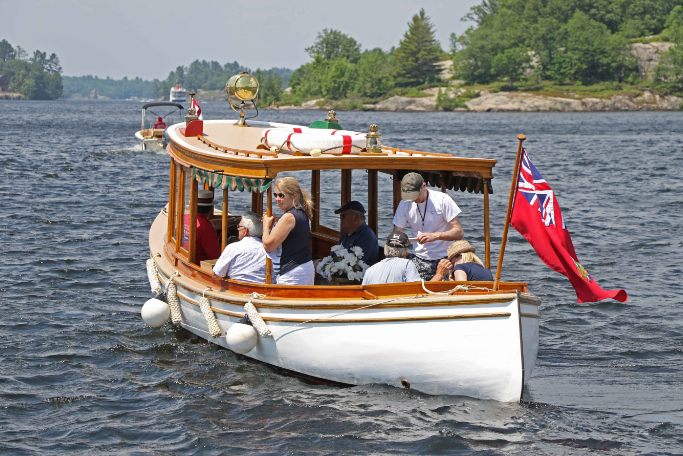 Wooden boat with several people cruising on a lake, with a red flag at the stern and trees along the shore.