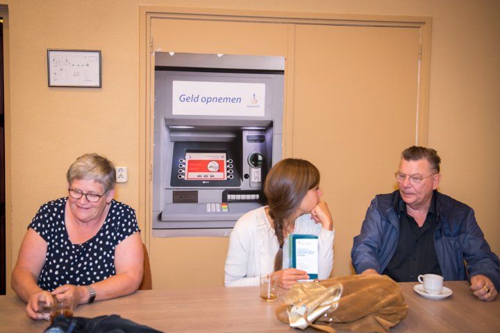 Drie mensen zitten aan een tafel voor een geldautomaat. De vrouw links glimlacht. Een andere vrouw en een man voeren een gesprek.