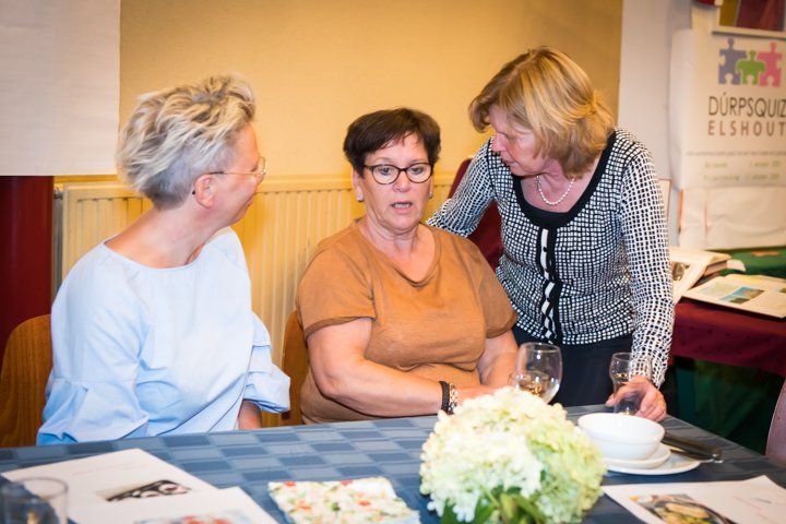 Drie vrouwen in gesprek aan een tafel; er zijn bloemen, eten en glazen aanwezig.
